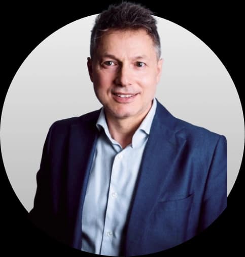 Zdjęcie uśmiechniętego Janusza Lisa - mężczyzny o krótkich, ciemnych włosach, ubranego w granatową marynarkę i błękitną koszulę, z otwartą postawą.